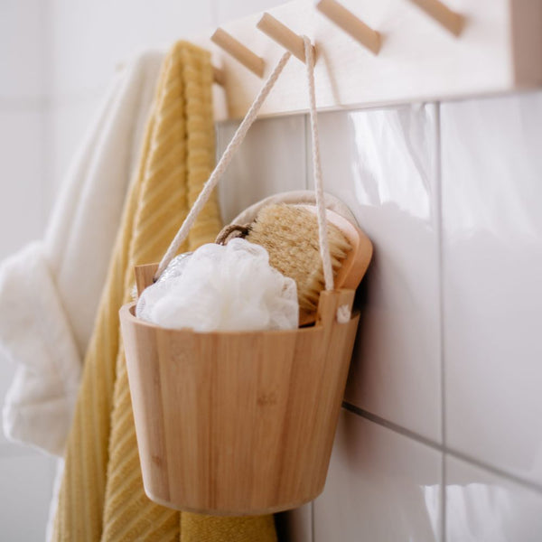 Ein Holzeimer mit Schlaufe hängt an einem Hakenbrett im Badezimmer, gefüllt mit einem Sisalhandschuh, einem Duschschwamm und einer Badebürste.